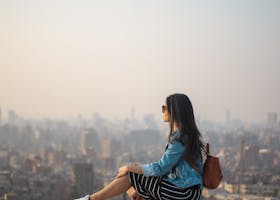 A young woman in casual attire sits on a rooftop terrace, viewing a sprawling cityscape in daylight.